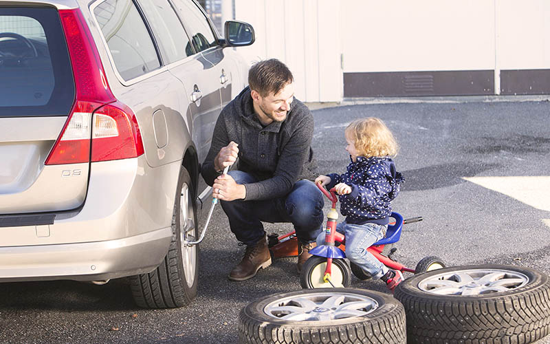 En man byter däck på en personbil som står parkerad på en uppfart. Intill sitter dottern på en trehjuling och tittar på. 